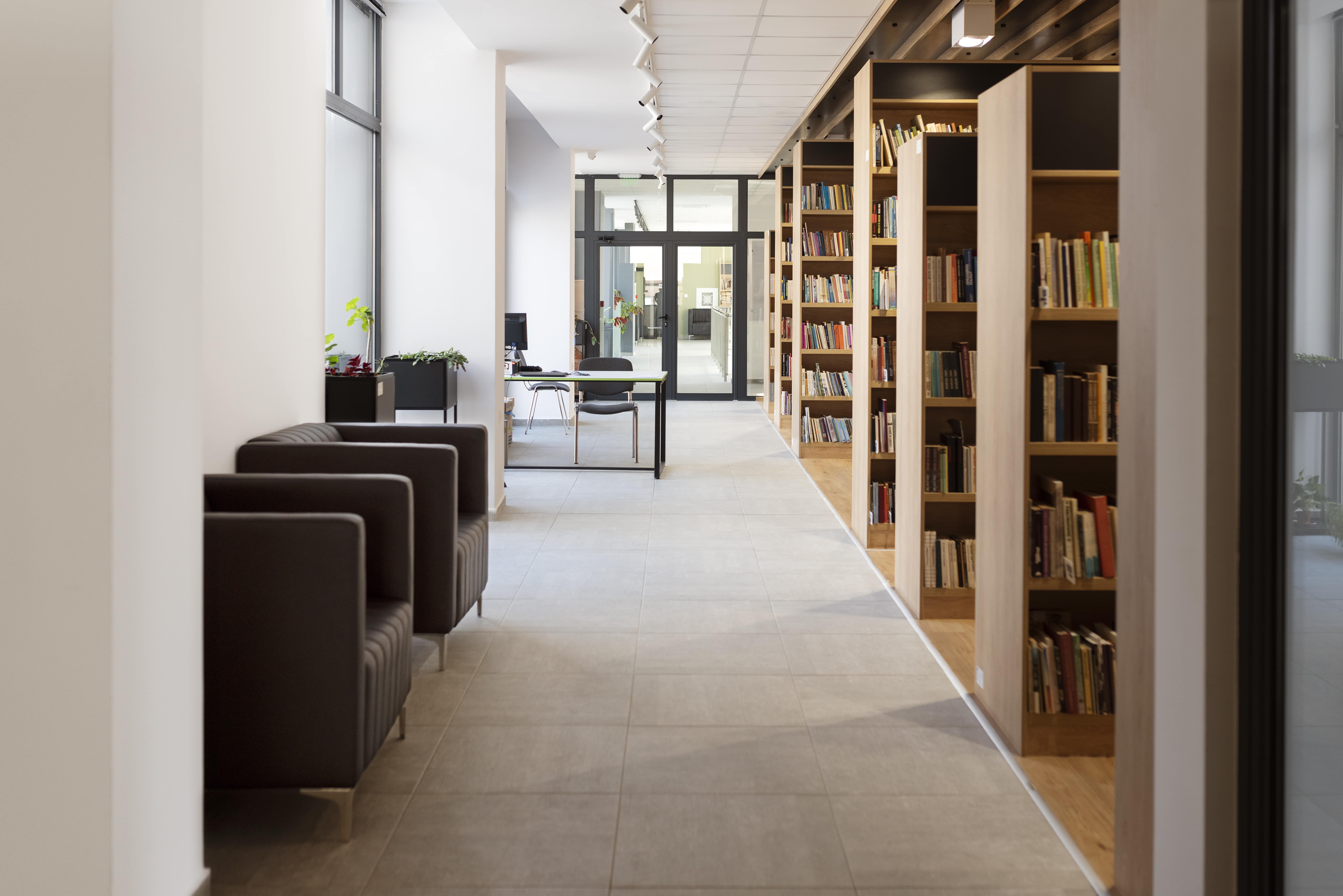 Interior de uma biblioteca moderna com estantes de livros, cadeiras para leitura e uma área de trabalho com mesa e cadeiras ao fundo.