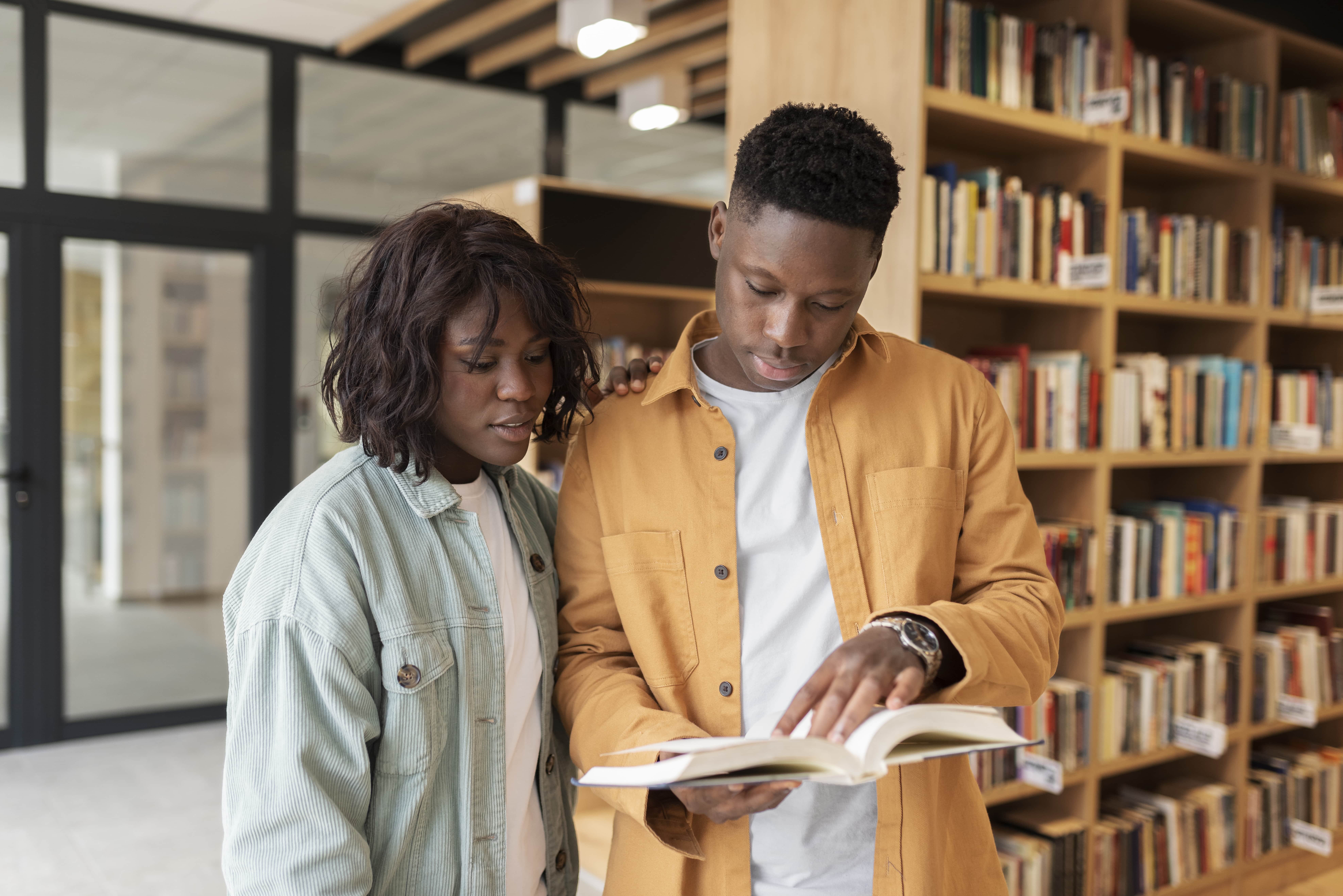 Casal jovem examinando um livro em uma biblioteca, com estantes de livros ao fundo.