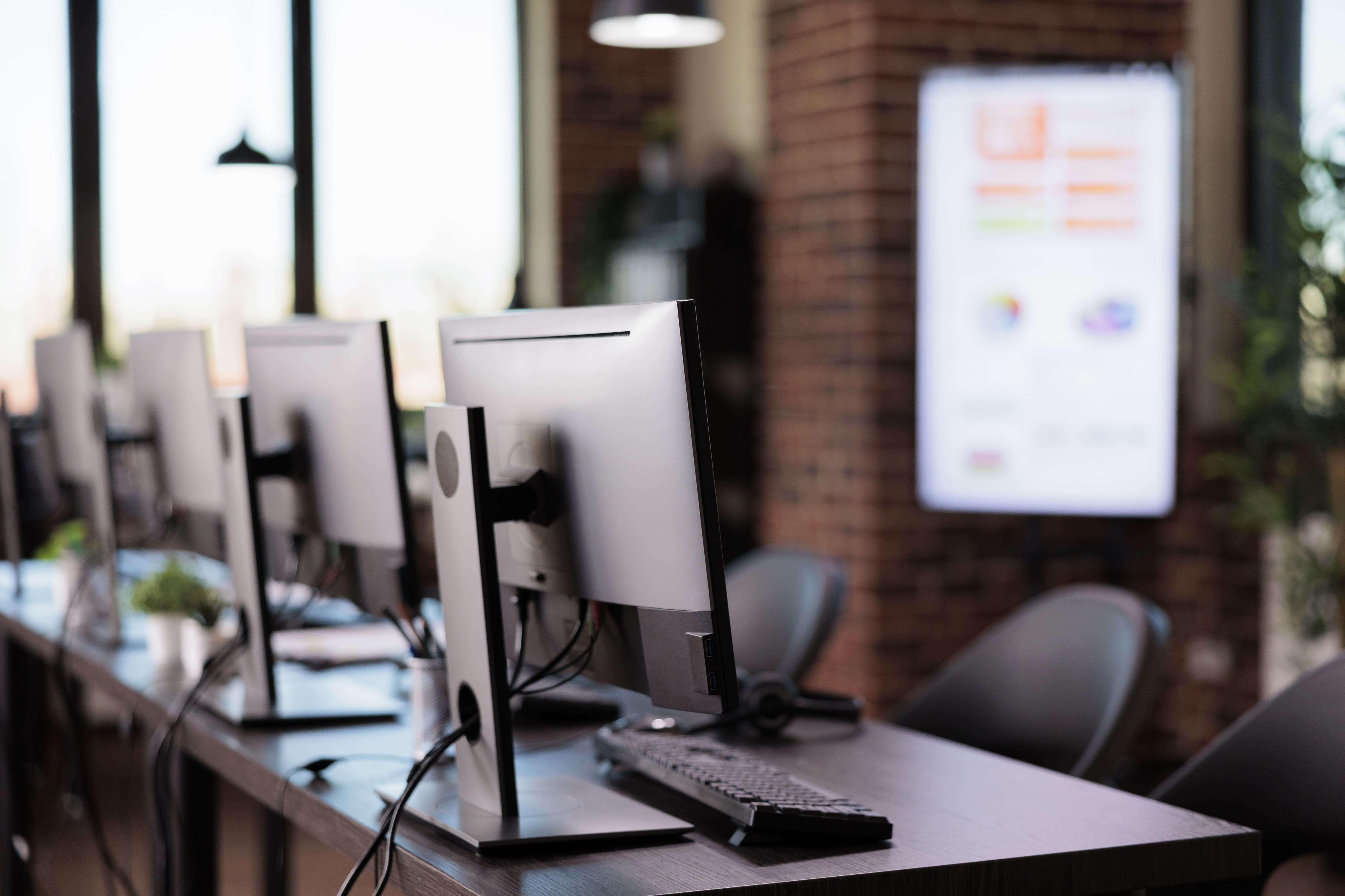 Fileira de monitores de computador em uma mesa em um ambiente moderno de escritório, com parede de tijolos e display digital ao fundo.