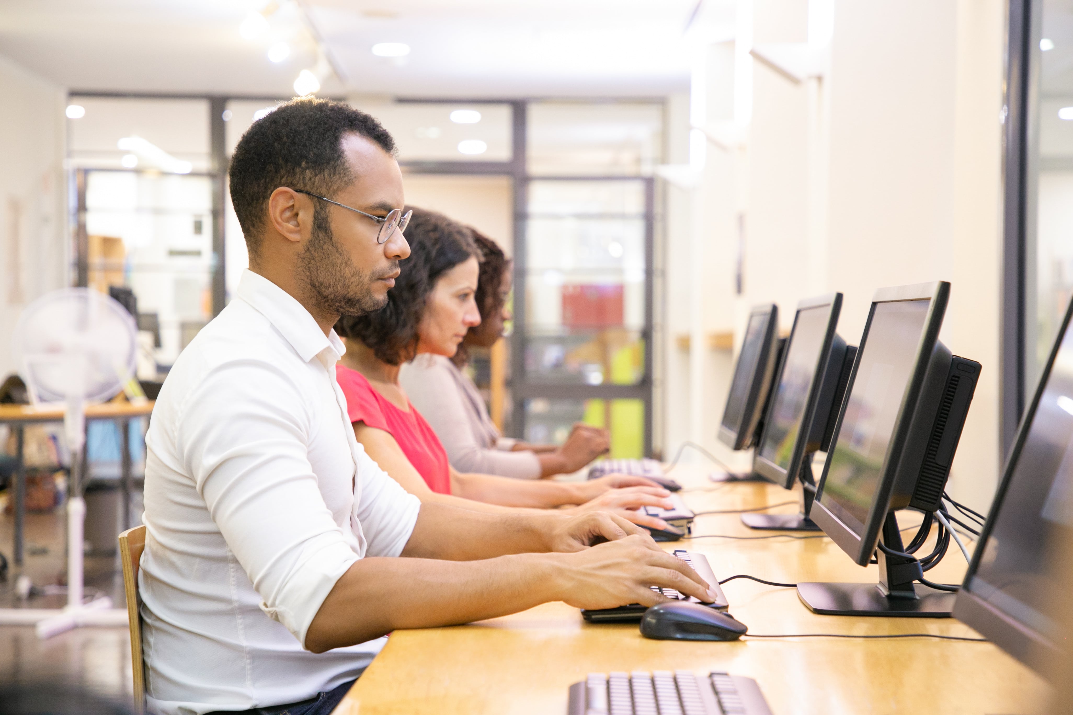 Três adultos trabalhando em computadores em uma sala de estudos ou laboratório de informática, sentados lado a lado e concentrados nas telas.